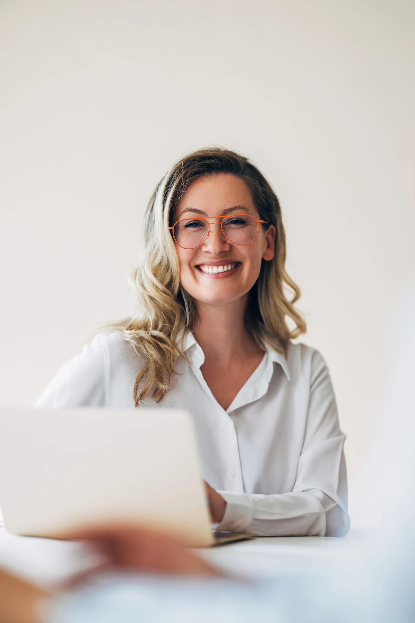 Close-up of a confident woman in an office setting, smiling while working on her laptop. She is wearing glasses and a professional white shirt.