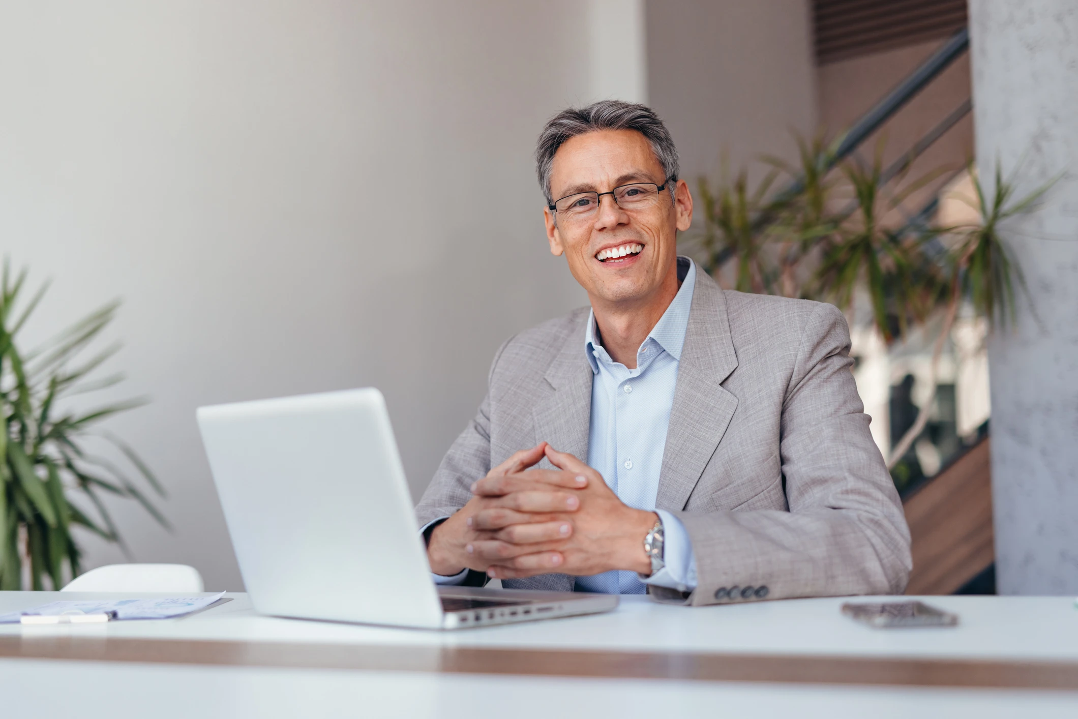 A middle-aged man in a light gray suit, sitting at a desk in a modern office environment. He is smiling warmly, with a laptop open, describing a sense of professionalism, confidence, and comfort.