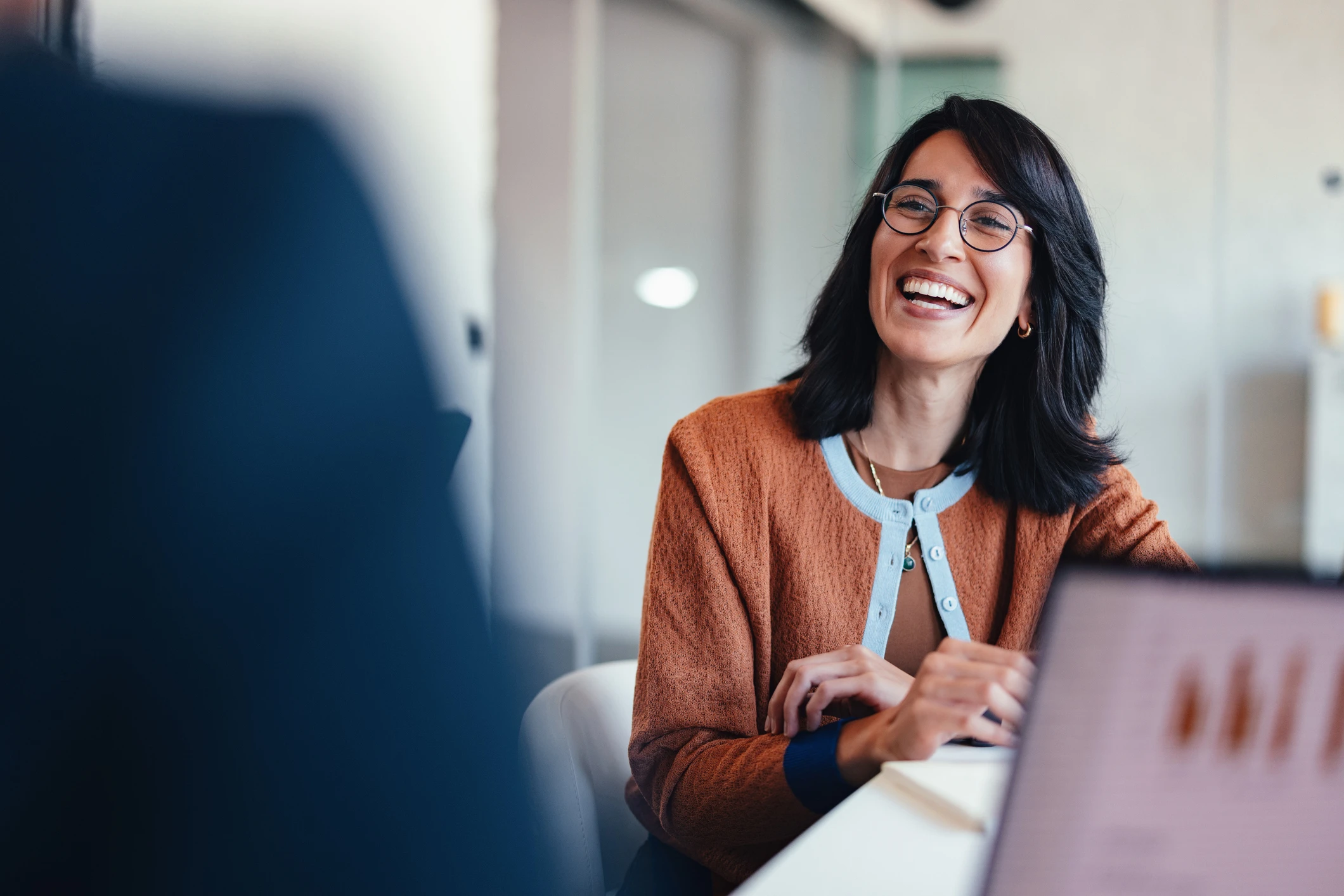 A cheerful female professional wearing round glasses sits at a modern office desk, chatting and sharing ideas. Her warm smile signals teamwork, collaboration, and a positive business atmosphere in a coworking or corporate setting.